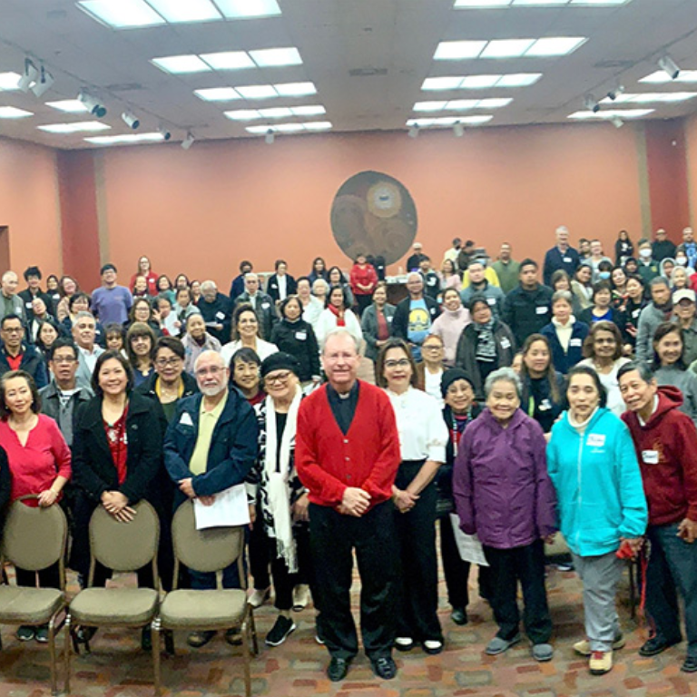 An image of the Filipino Catholic Charismatic community gathered in a meeting hall and listening to a Priest talking on stage.
